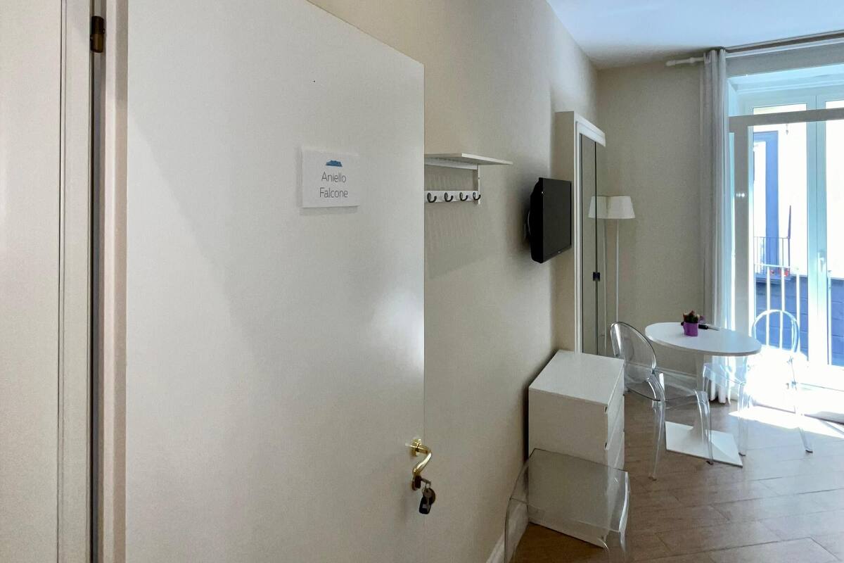 A hotel room with a door labeled "Anieleo Falcone," a small white table with chairs, a TV mounted on the wall, and a window with a view of the outside.