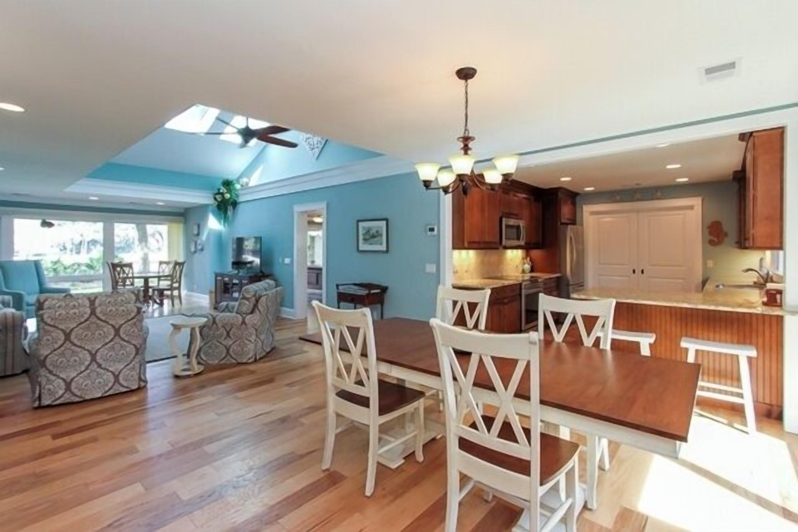 A spacious living area with a wooden dining table and white chairs, a kitchen with wooden cabinets, and a ceiling fan.
