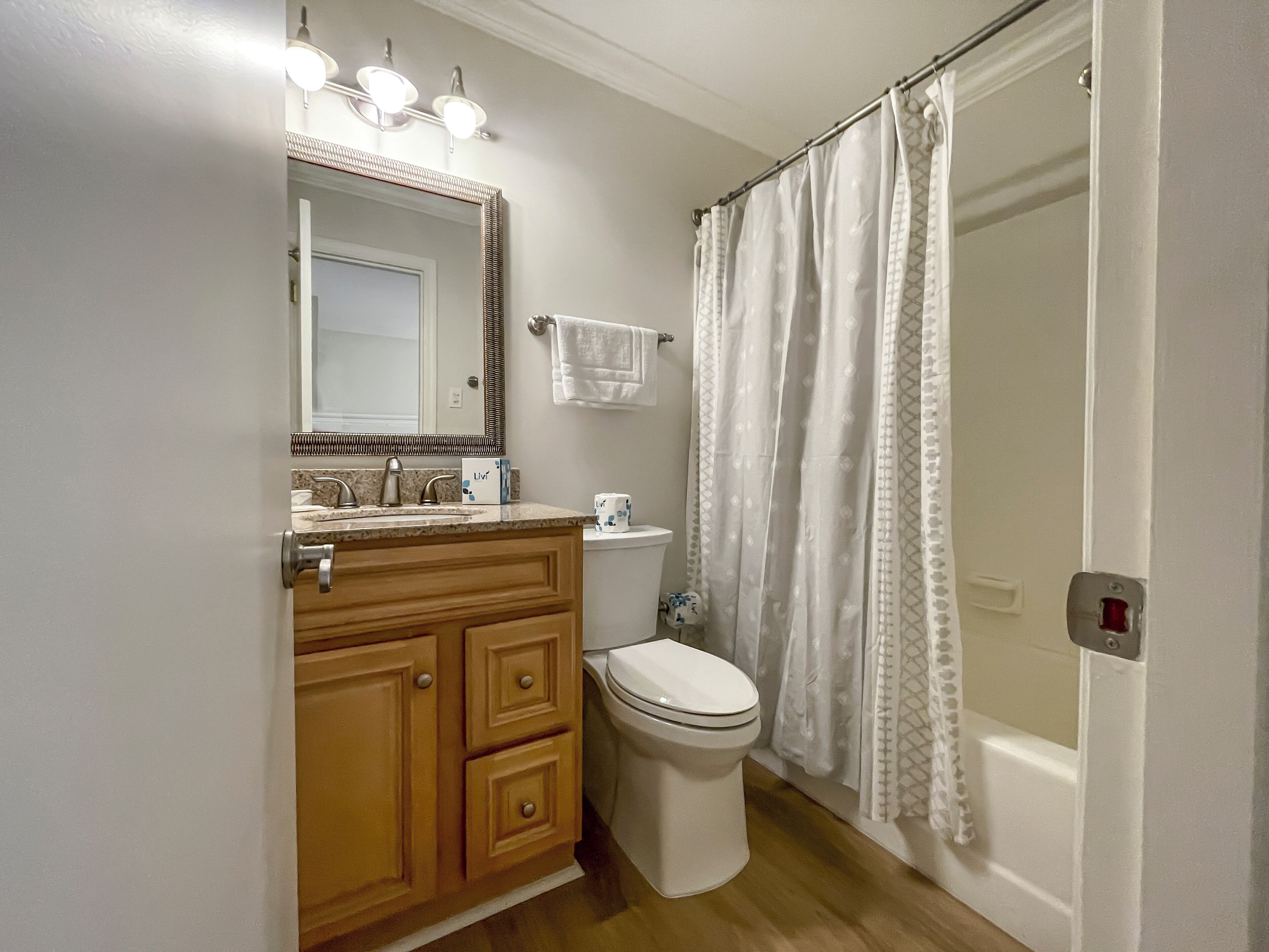 Une salle de bain avec un meuble-lavabo en bois, des toilettes blanches, un rideau de douche et un miroir avec des lumières au-dessus.