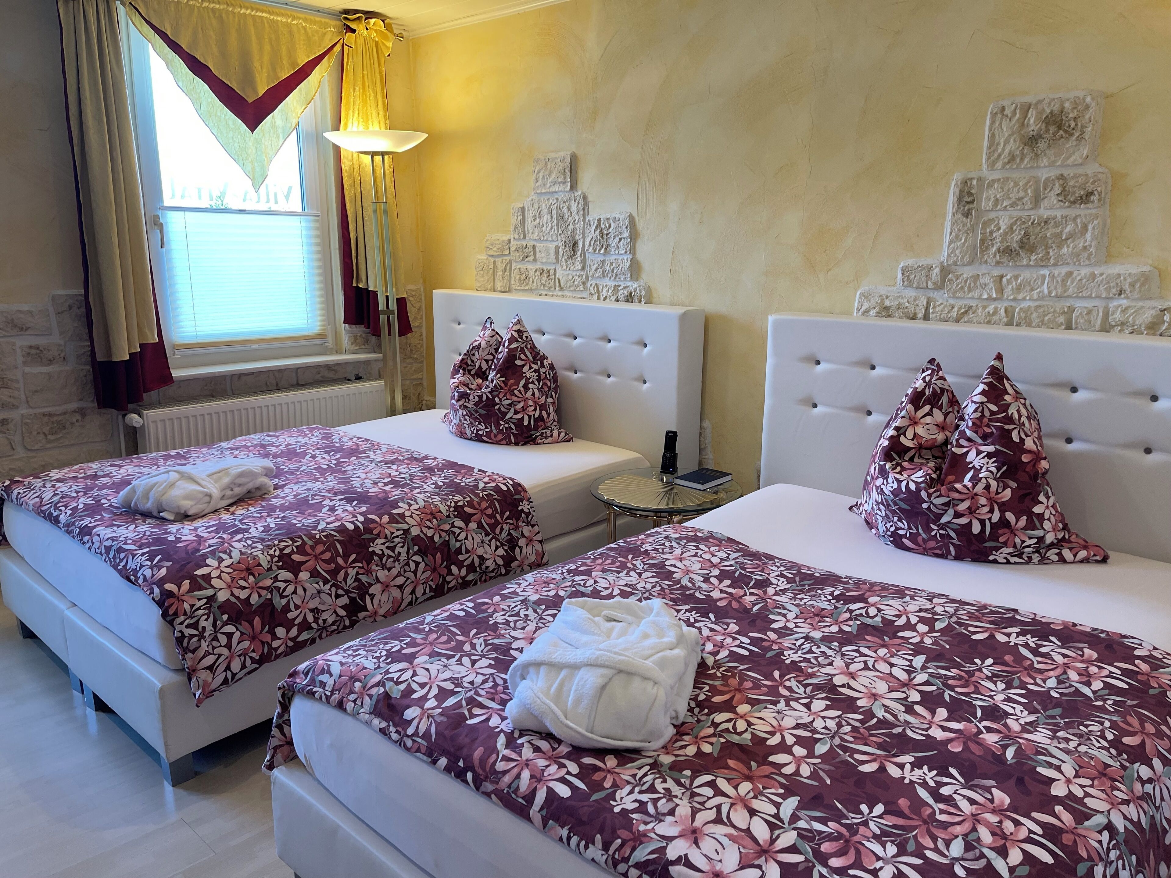 A hotel room with two beds, each with a floral bedspread and a white headboard. There is a small round table between the beds with a remote control and a book on it. The room has a stone wall and a window with curtains.