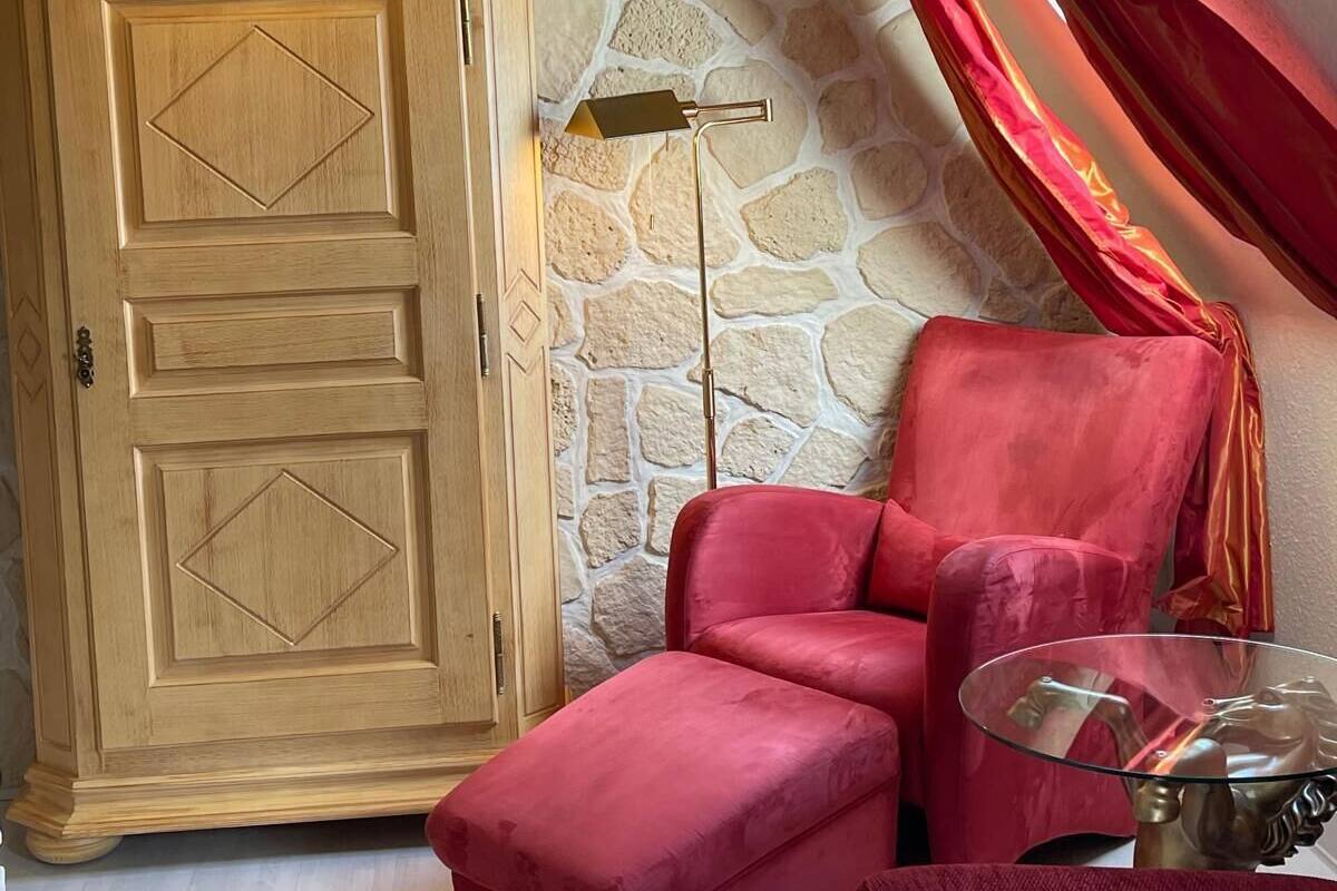 Una habitación con un armario de madera, un sillón rojo, una lámpara de pie y una pared de piedra.