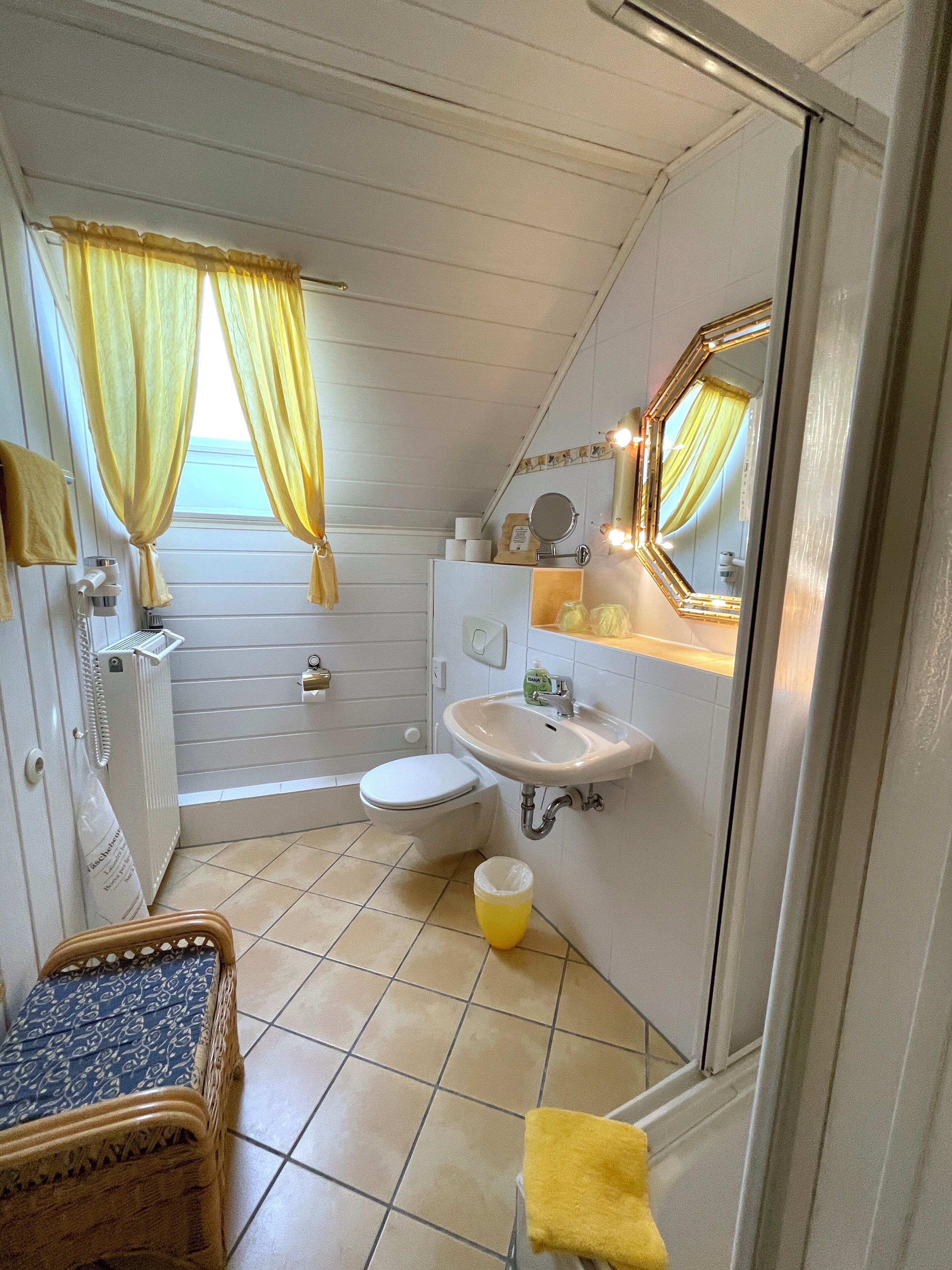 A compact, well-lit bathroom with a toilet, sink, and mirror. Yellow curtains and a small stool are visible.