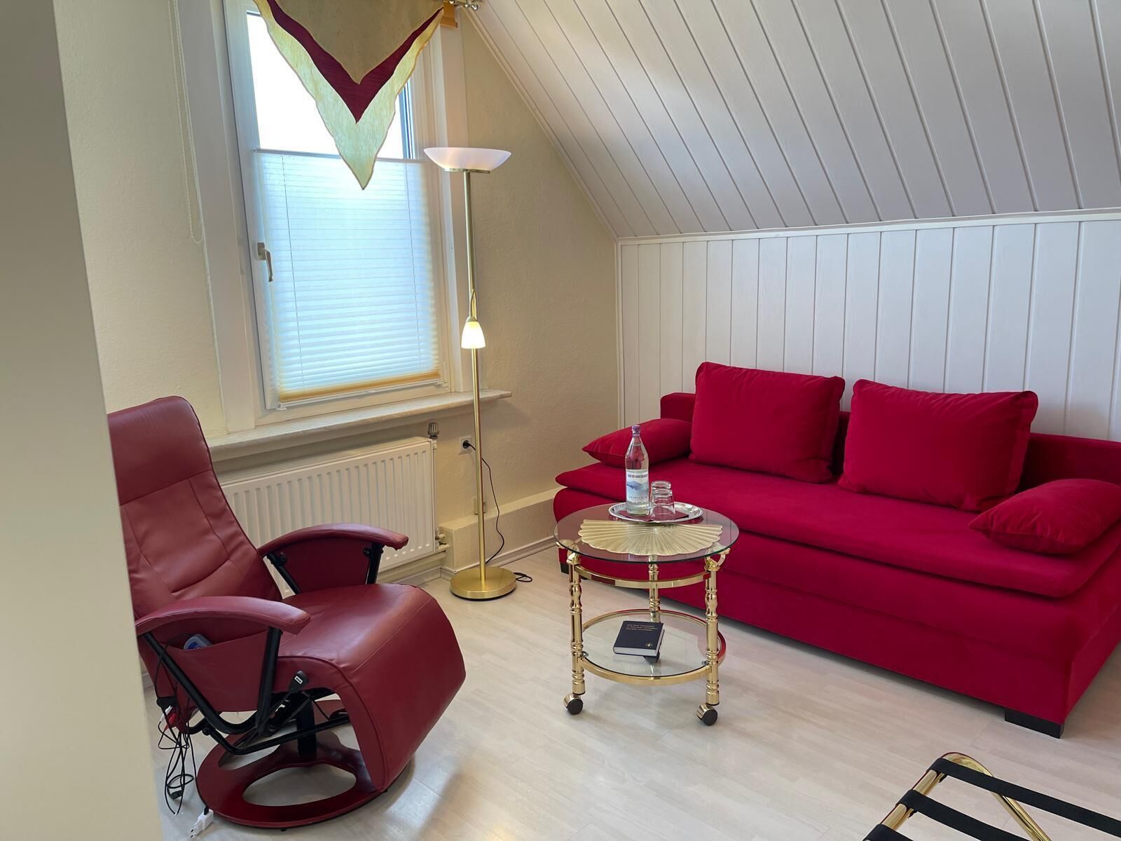 A red leather chair, a red sofa, a small round table with a book, and a floor lamp in a room with white walls and a sloped ceiling.