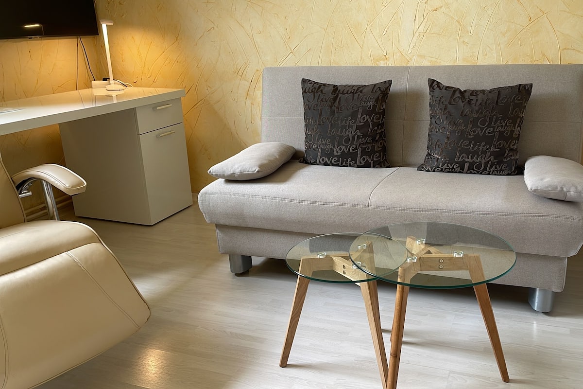 Una sala de estar moderna con un sofá gris, dos almohadones estampados, una mesa de centro de vidrio y un escritorio blanco con una lámpara.