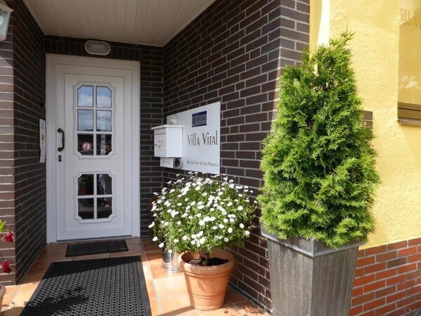 Un bâtiment en briques avec une porte blanche, une enseigne indiquant « Villa Vital » et deux plantes en pot.