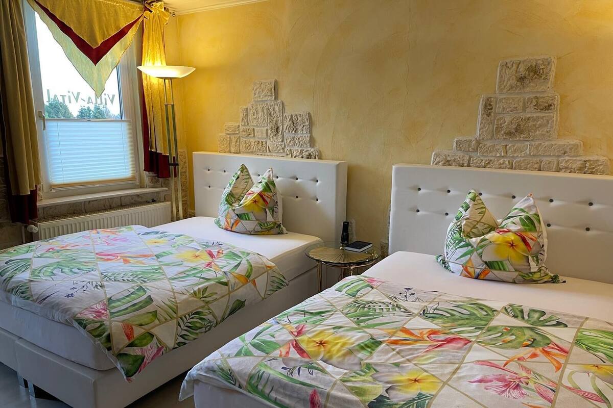 Habitación de hotel con dos camas, cada una con ropa de cama y almohadas a juego de estampado floral. Hay una pequeña mesa redonda entre las camas, y una decoración de pared de piedra sobre ellas.