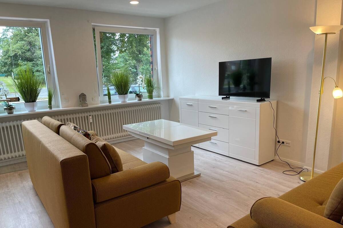 Un salón moderno con un sofá, una mesa de centro y un televisor sobre un mueble blanco. Hay ventanas con plantas y vistas a zonas verdes.