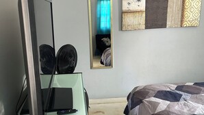 Ett sovrum med en säng, ett skrivbord med en dator och två fläktar, en spegel och ett konstverk på väggen.