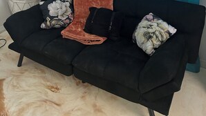 En svart soffa med en brun pläd och blommiga kuddar, placerad framför en blå gardin.