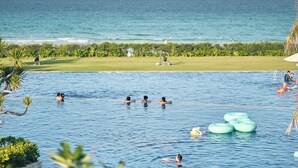 A swimming pool with people swimming and floating, a golf course in the background, and a view of the ocean. - Ocean Sun Pool Villas Resort Cam Ranh (Cam Lam)