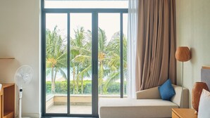A hotel room with a bed, a sitting area with a sofa and a blue pillow, a standing fan, and a view of palm trees through the window. - Ocean Sun Pool Villas Resort Cam Ranh (Cam Lam)