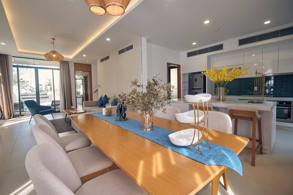 A modern living room with a long wooden dining table, comfortable seating, a kitchen area, and a large window providing natural light.