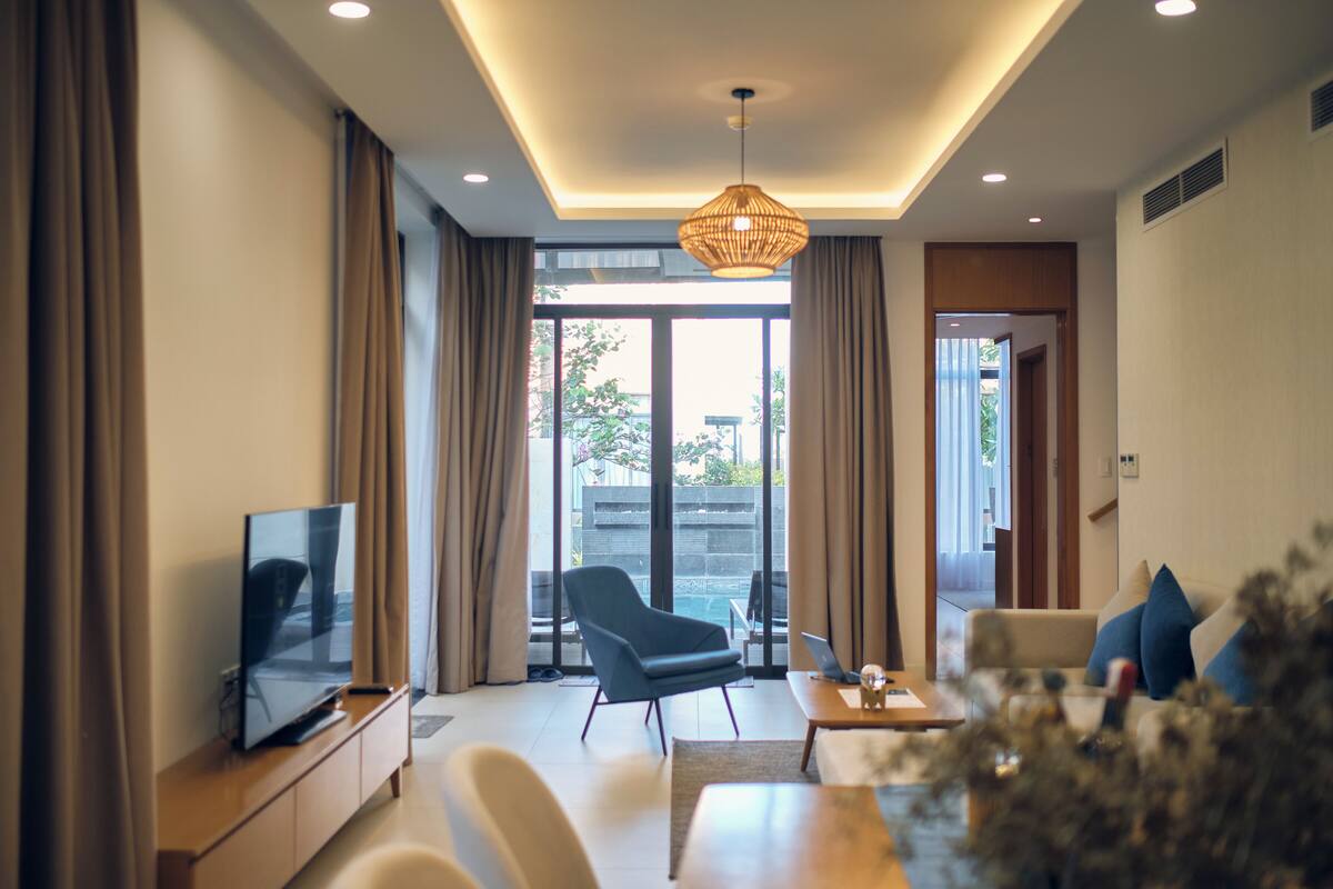 A modern living room with a flat-screen TV, a sofa, a chair, and a dining table. There is a large window with curtains and a view of the outdoors.