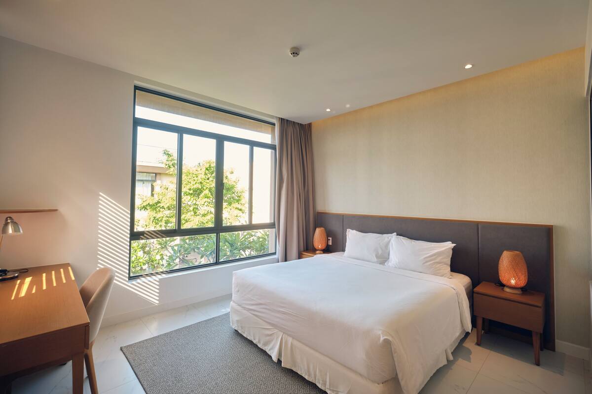 A modern hotel room with a large bed, bedside lamps, a desk, and a window offering a view of greenery.