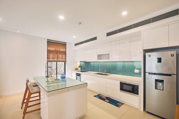 A modern kitchen with white cabinetry, a glass island, stainless steel appliances, and a window with blinds. - Ocean Sun Pool Villas Resort Cam Ranh (Cam Lam)