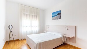 A bedroom with a wooden floor, a bed with white bedding, a bedside table, a wall-mounted shelf, a picture on the wall, and a window with sheer curtains. - DREAM SEA APARTMENTS - 1 BEDROOM (El Ejido)