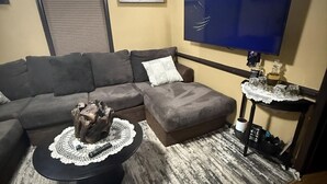 A living room with a sectional sofa, a coffee table with a decorative centerpiece, a flat-screen TV mounted on the wall, and a small side table with a lace doily.