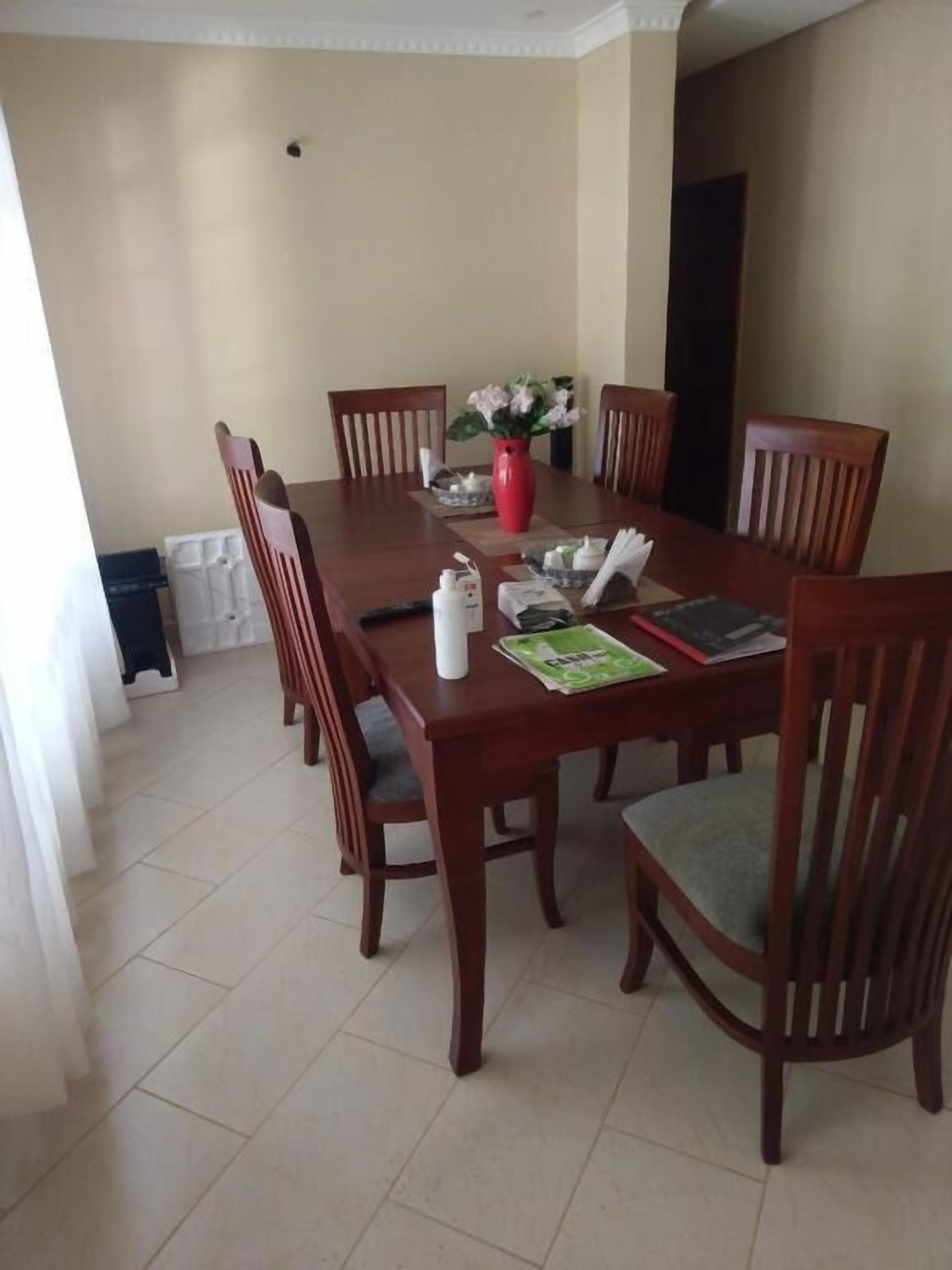 A dining area with a rectangular wooden table surrounded by six wooden chairs with green cushions. A red vase with flowers is placed on the table.