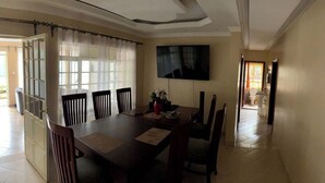 A dining area with a large table, chairs, a TV mounted on the wall, and a view of the outside through a window with curtains. - GreenHaven Siaya Cottages (Siaya)