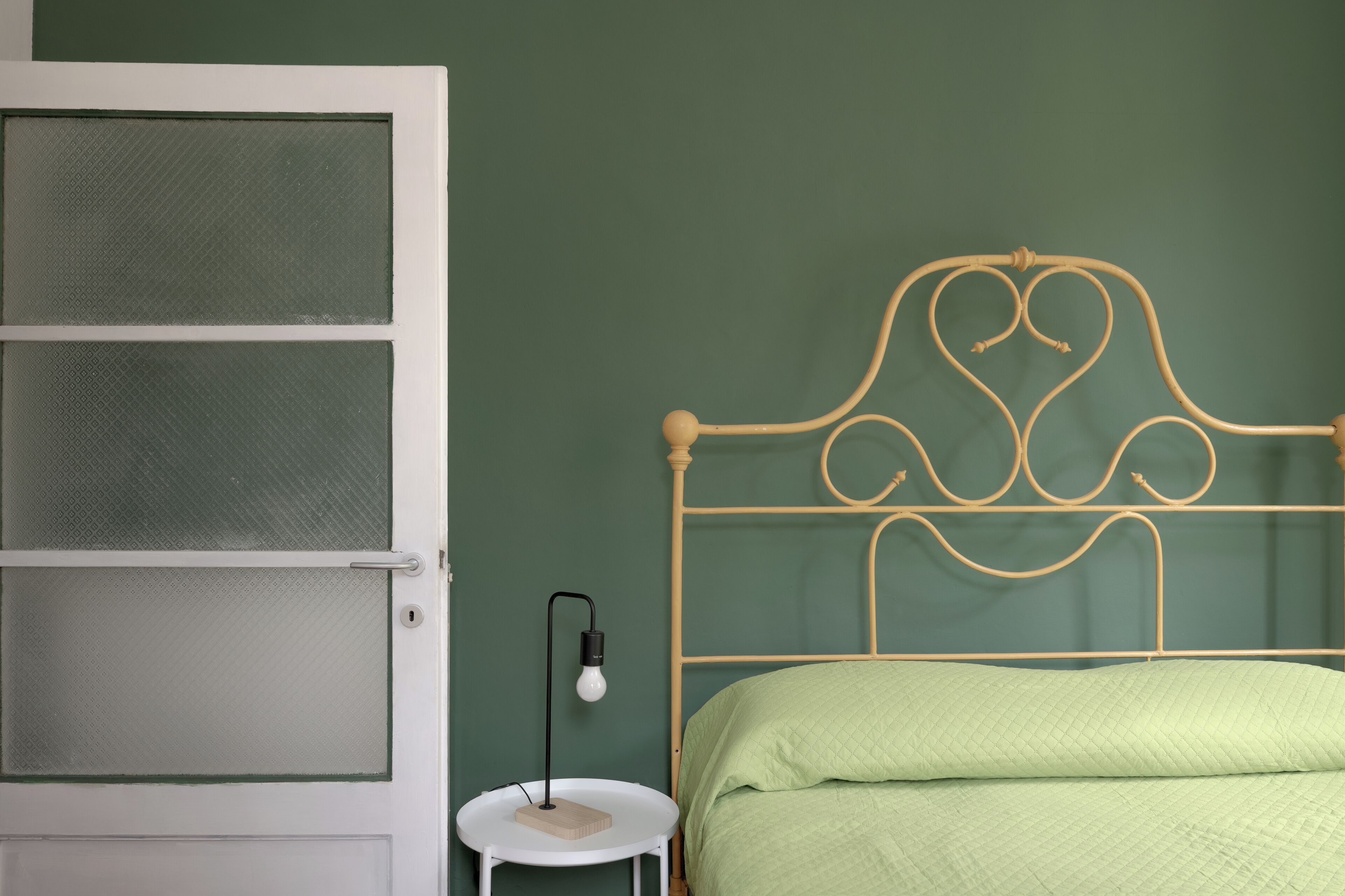 Une pièce avec un lit dont le cadre est en métal, une table ronde blanche avec une lampe et une porte munie d’une moustiquaire.