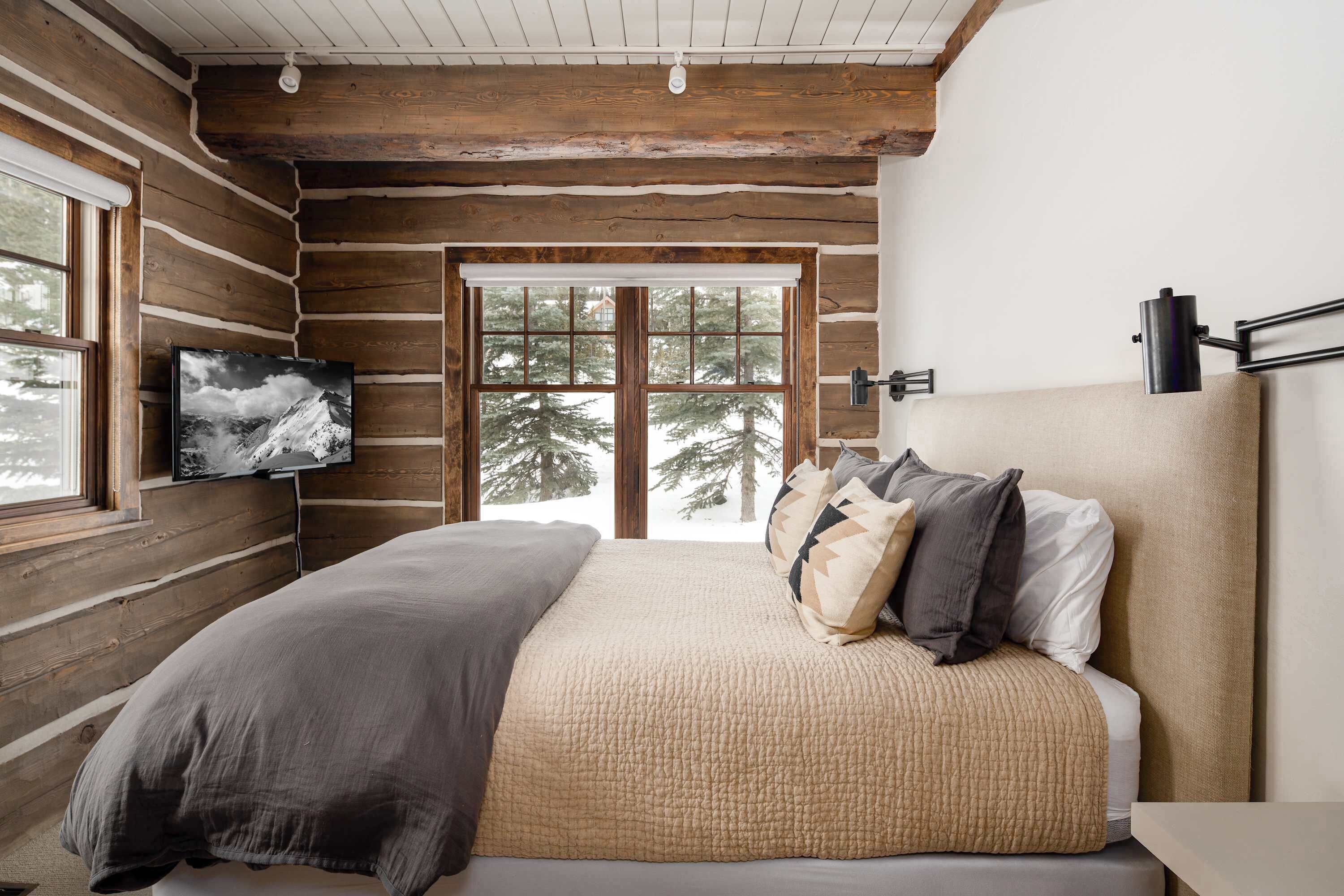 Ein Schlafzimmer in einer Blockhütte mit einem Bett, einem an der Wand befestigten Fernseher und Blick auf Bäume durch ein großes Fenster.