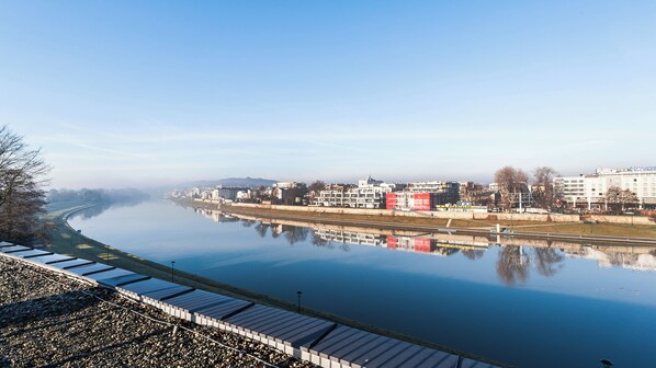 A river with buildings on the opposite bank, a clear sky, and a calm water surface reflecting the buildings. - Apartamenty Sun & Snow Riva Verona (Kraków)