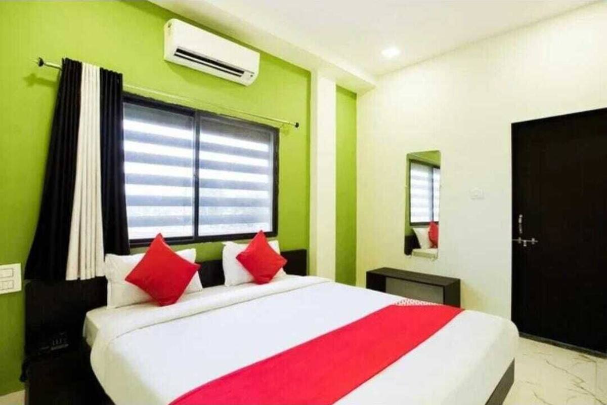 A hotel room with a bed, a window with curtains, an air conditioner, a bedside table, and a door.