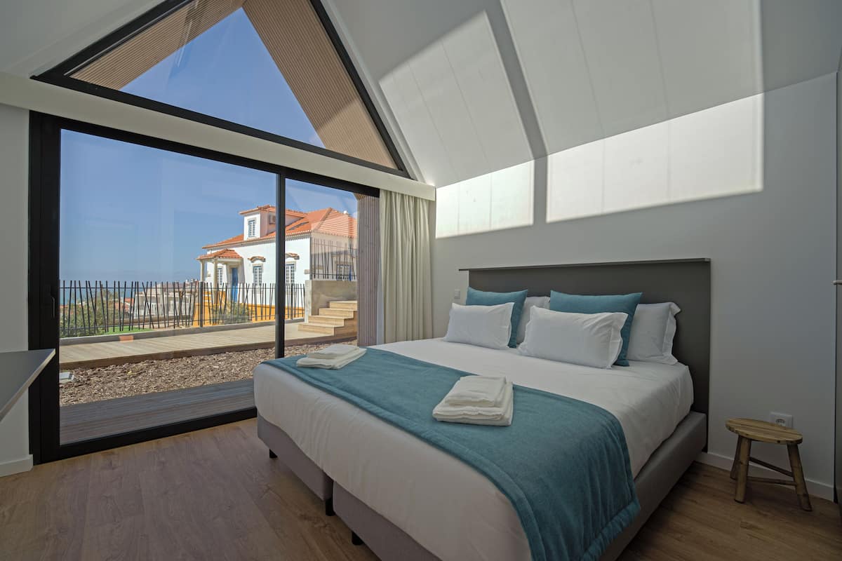 Ein modernes Schlafzimmer mit einem großen Bett, Blick auf ein Gebäude durch eine Glasdoor und Holzboden.