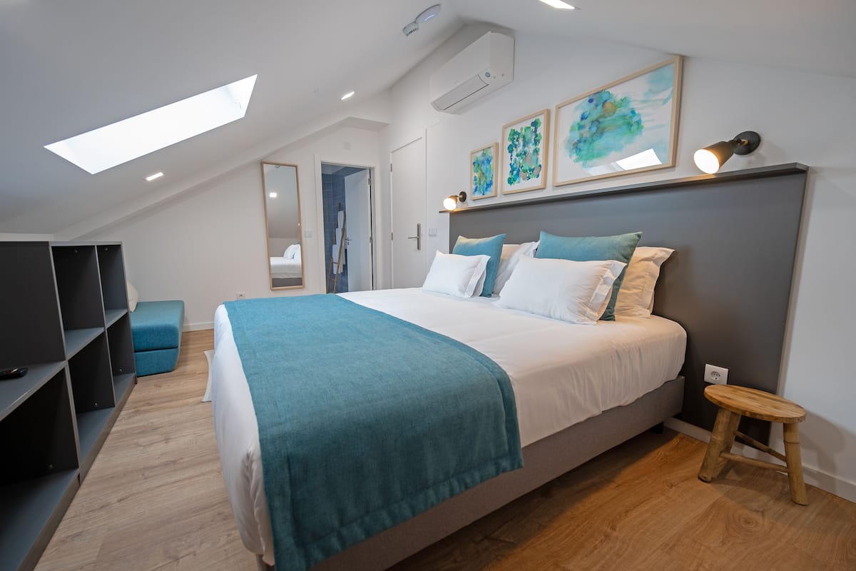 Ein modernes Schlafzimmer mit Oberlichter, einem Bett mit Türkis-Decke, einem kleinen Hocker und einem Fernseher auf einem Regal.
