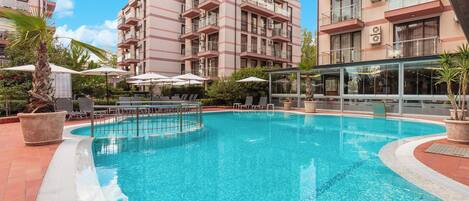 A swimming pool with lounge chairs and umbrellas, surrounded by multi-story buildings with balconies.
