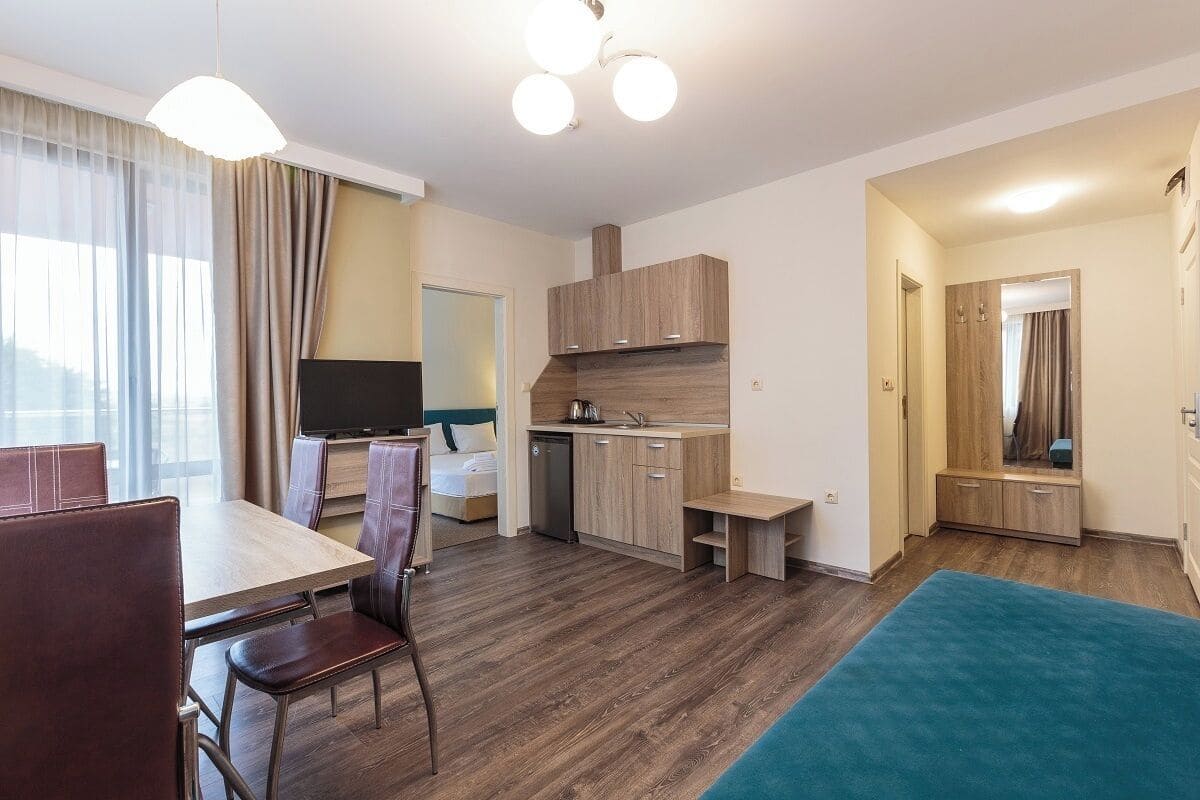 A modern hotel room with a dining area, kitchenette, and a large window with curtains.