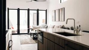 A modern kitchen with a large island, a built-in oven, and a sink. There is a dining area with a sofa and a coffee table.