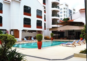 Una piscina con sillas de descanso, una zona de estar cubierta y un edificio de varios pisos con balcones.