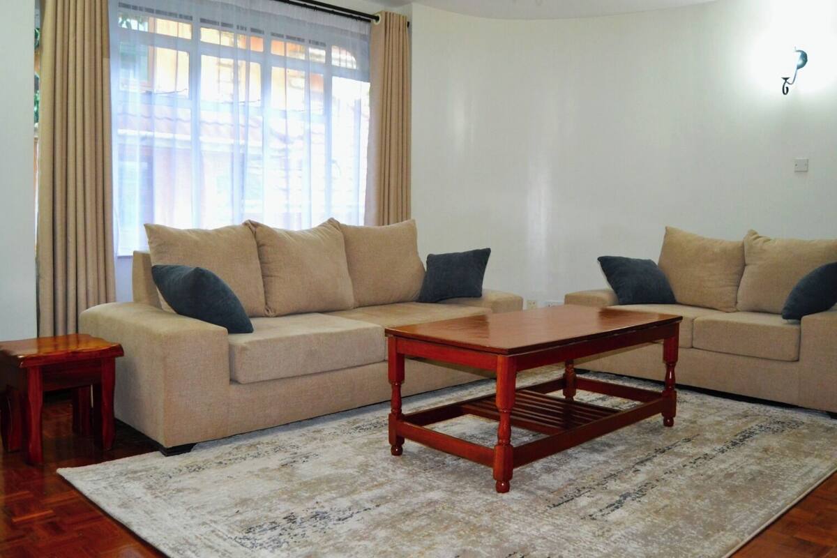 Een woonkamer met twee beige banken, een houten salontafel, een klein houten bijzettafeltje en een kamerkleed met een patroon.