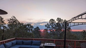 A deck with a sofa, table, and chairs, overlooking a forested landscape at sunset.