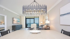 Uma sala de jantar moderna com uma mesa grande, cadeiras, um lustre decorativo e vista para o oceano através de amplas janelas.