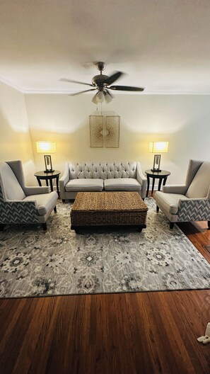 A living room with a grey sofa, two armchairs, a coffee table, a patterned rug, a ceiling fan, and two lamps.