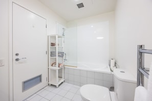 Un baño con inodoro blanco, bañera, ducha y estante blanco con toallas.