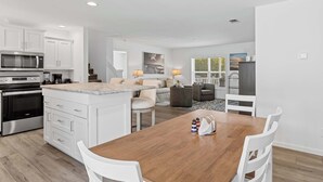 Een moderne keuken met witte kasten, een ingebouwde magnetron en een oven. Er is een eethoek met een houten tafel en witte stoelen.