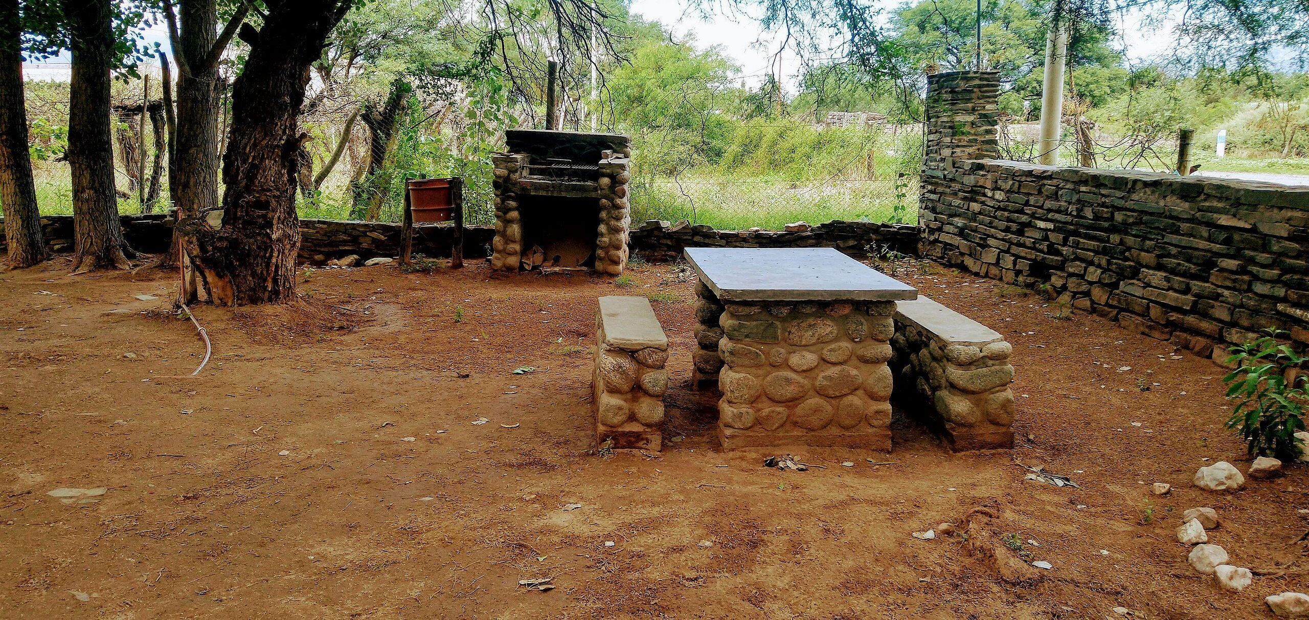 An outdoor stone barbecue area with a table and benches, surrounded by trees and a stone wall.