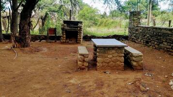 An outdoor stone barbecue area with a table and benches, surrounded by trees and a stone wall.