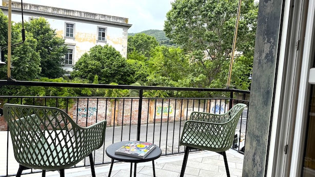 A balcony with two green chairs and a small table, overlooking a street with graffiti on the wall and trees in the background. - Kennedy 2 - One Bedroom Apartment, Sleeps 3 (Albenga)