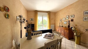 A dining area with a table, chairs, a vase with flowers, a window with curtains, and a cabinet with framed pictures. - Kennedy 2 - One Bedroom Apartment, Sleeps 3 (Albenga)