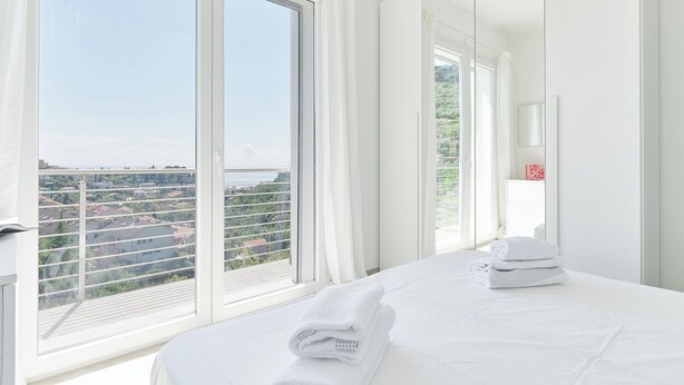 A hotel room with a large window, a bed with white linens, and a view of a cityscape. - The Italian Lady - Unit 1 (Alassio)