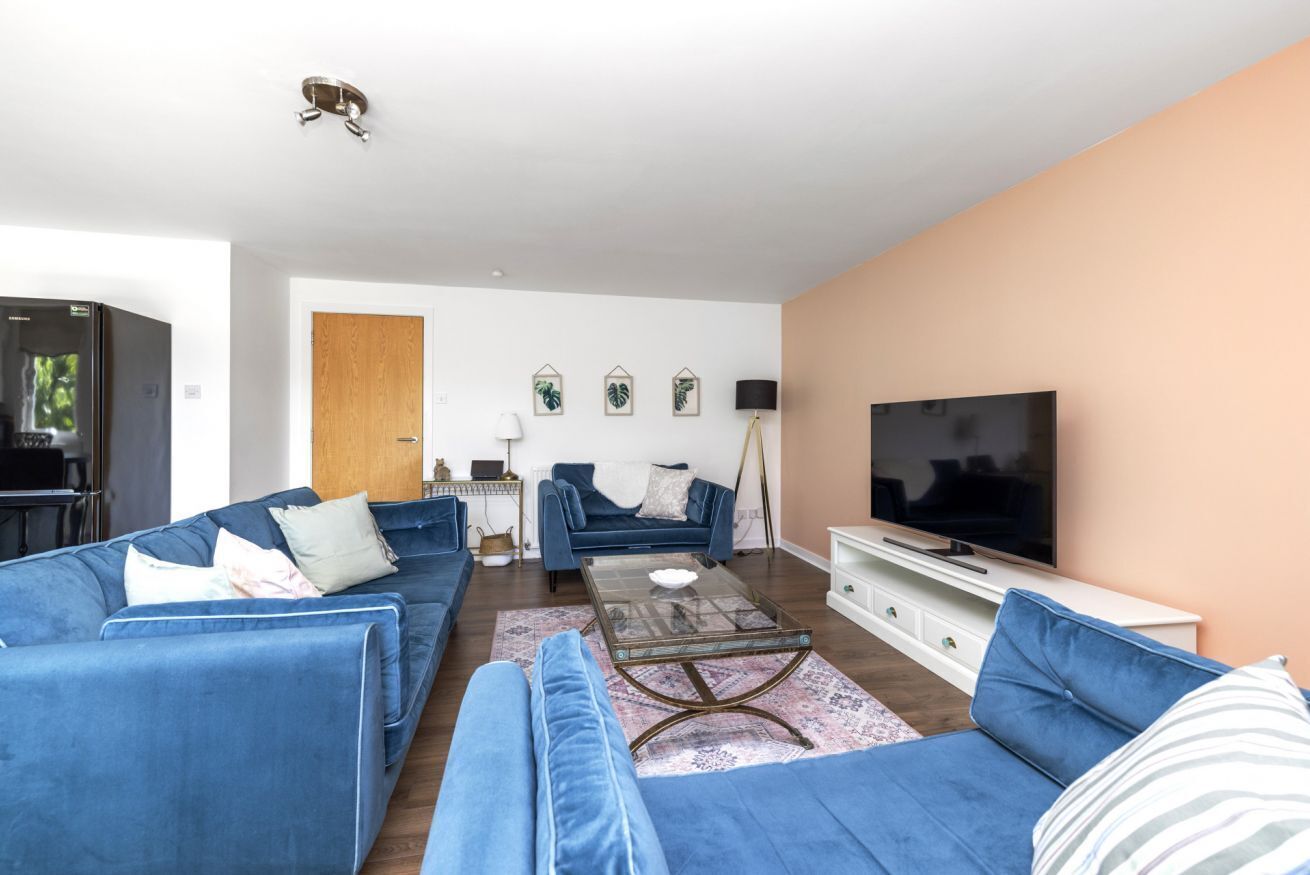 Een moderne woonkamer met een blauwe zithoek, een glazen salontafel, een flatscreen televisie en een houten kast.