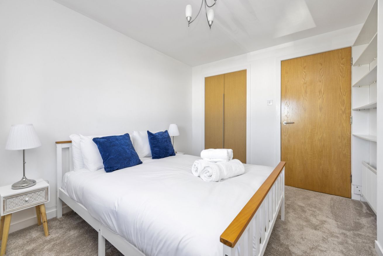 A bedroom with a bed, a nightstand, a lamp, a wooden door, and a white wardrobe.