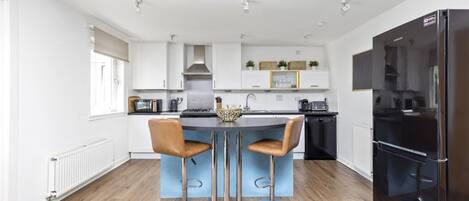 A modern kitchen with a central island, two bar stools, a black refrigerator, and white cabinetry.