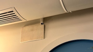Ein Zimmerwinkel mit weißer Wand, einer blauen Tür und einem weißen Lüftungsgitter an der Decke.