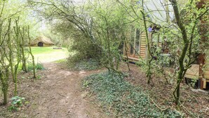 A rustic wooden cabin surrounded by dense greenery and a dirt path leading to a thatched-roof structure in the background. - Brandon House Farm Yurt (Norwich)