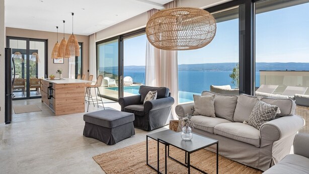 A modern living room with a large sofa, a coffee table, and a dining area with a bar. The room has large windows offering a view of the sea. - Ocean View Villa: Your Dream Getaway (Jesenice, Dugi Rat)